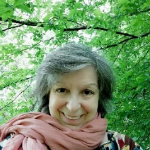 Porträt einer lächelnden Frau mit lockigem grauen Haar und einem rosa Schal vor grünem Laub.