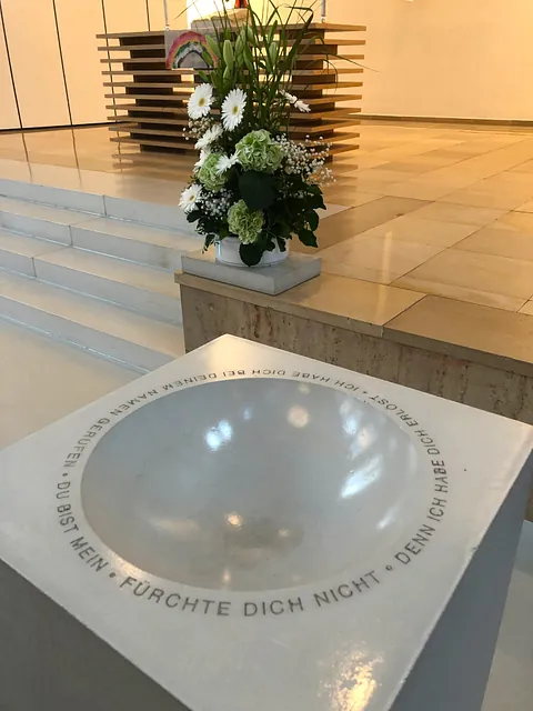 Taufbecken aus Stein mit Inschrift vor einem Altar mit Blumen.