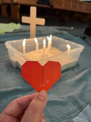 Brennende Kerzen in Sand, mit einem roten Papierherz davor.