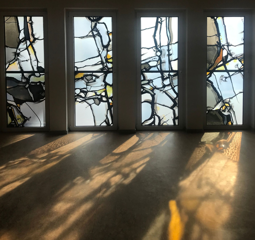 Vier große Fenster mit abstrakten Mustern werfen Sonnenlicht Schatten auf den Boden.