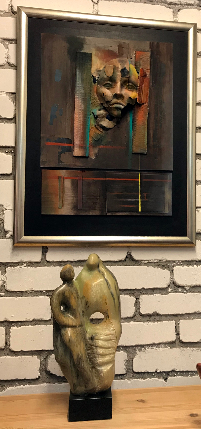 Gerahmtes, abstraktes Kunstwerk über einer Skulptur auf einem Regal vor einer Ziegelwand.